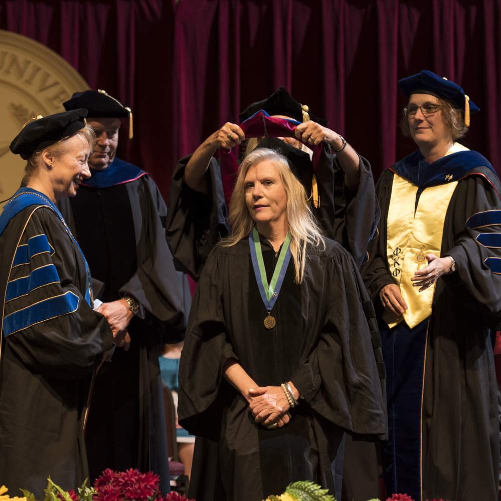 During a Summer 2016 commencement ceremony, a Doctoral candidate is hooded by her supervising professors; Dr. Golato, Dean of the Graduate College; and President Denise M Trauth.