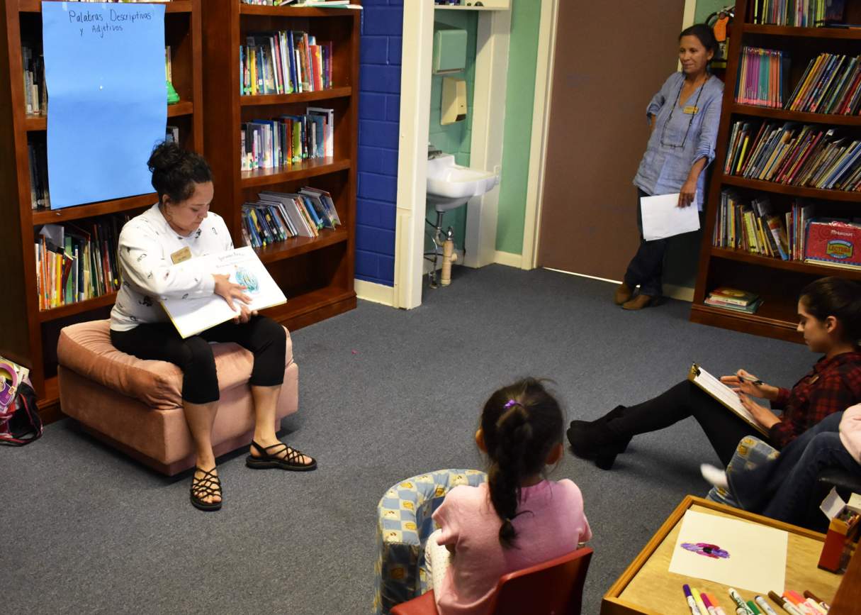 Jacqueline Tellez reads a bilingual book to SMCISD students while her fellow student Yamilex Yamilex Hernández takes notes. Dr. Luz Murillo observes from the doorway. 