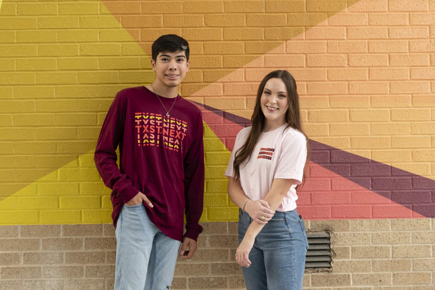 Two students in front of a colorful mural modeling TXST Next shirts