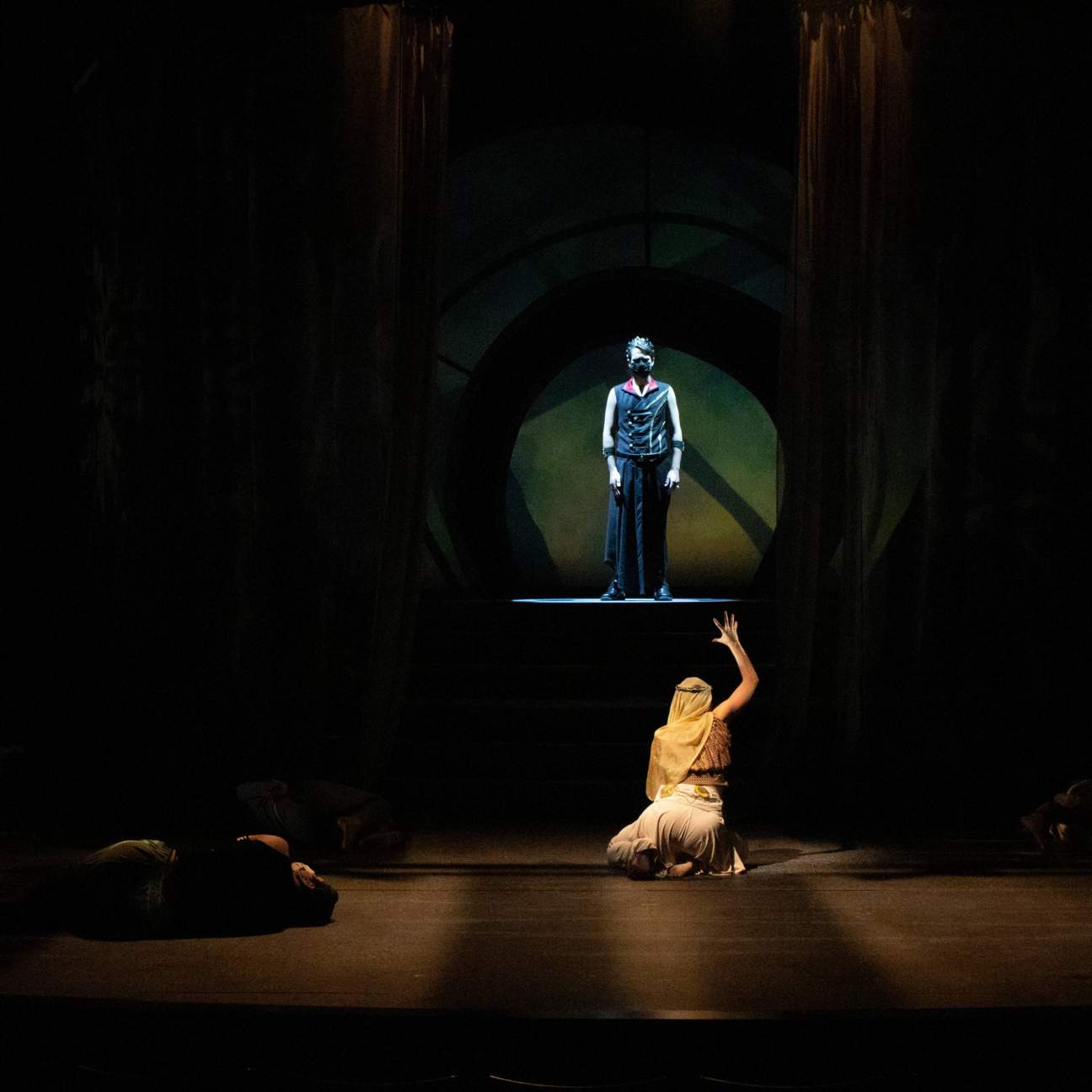 The King stands on a raised platform with a light shining on him from above. A female chorus member crawls to him, reaching her arms out to him.