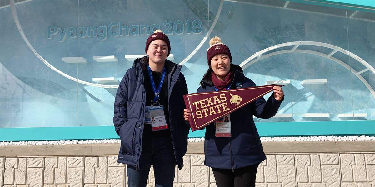 two texas state students hold up a texas state banner in front of the Pyeongchang 2018 Olympics