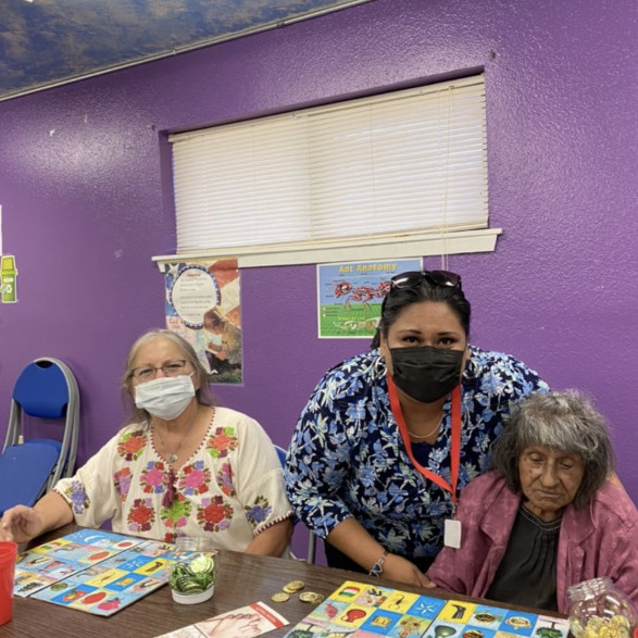 Two women (one masked, another unmasked) seated and playing Bingo, one woman (masked) standing behind one of the two women.