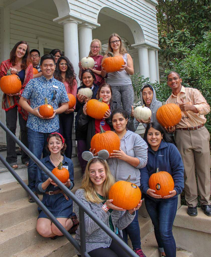 A group of students and advisors smile, holding pumpkins, in front of the Thornton International House