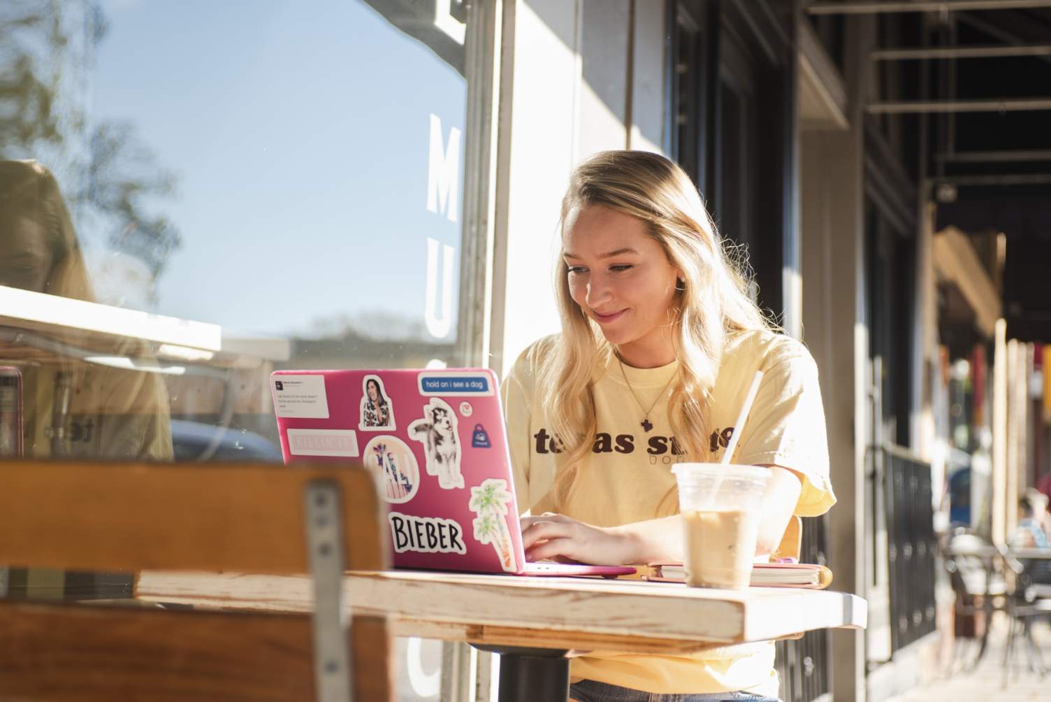 A young woman sitting at an outdoor cafe table working on a computer with an iced coffee next to it on the table.
