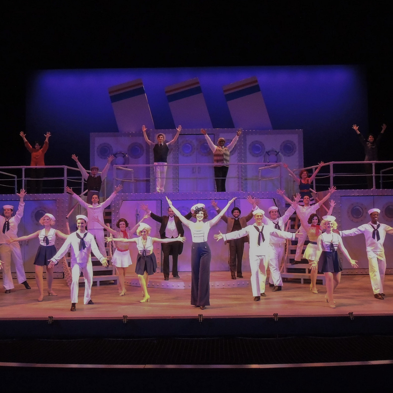 full cast on stage, dancing with arms in the air. 