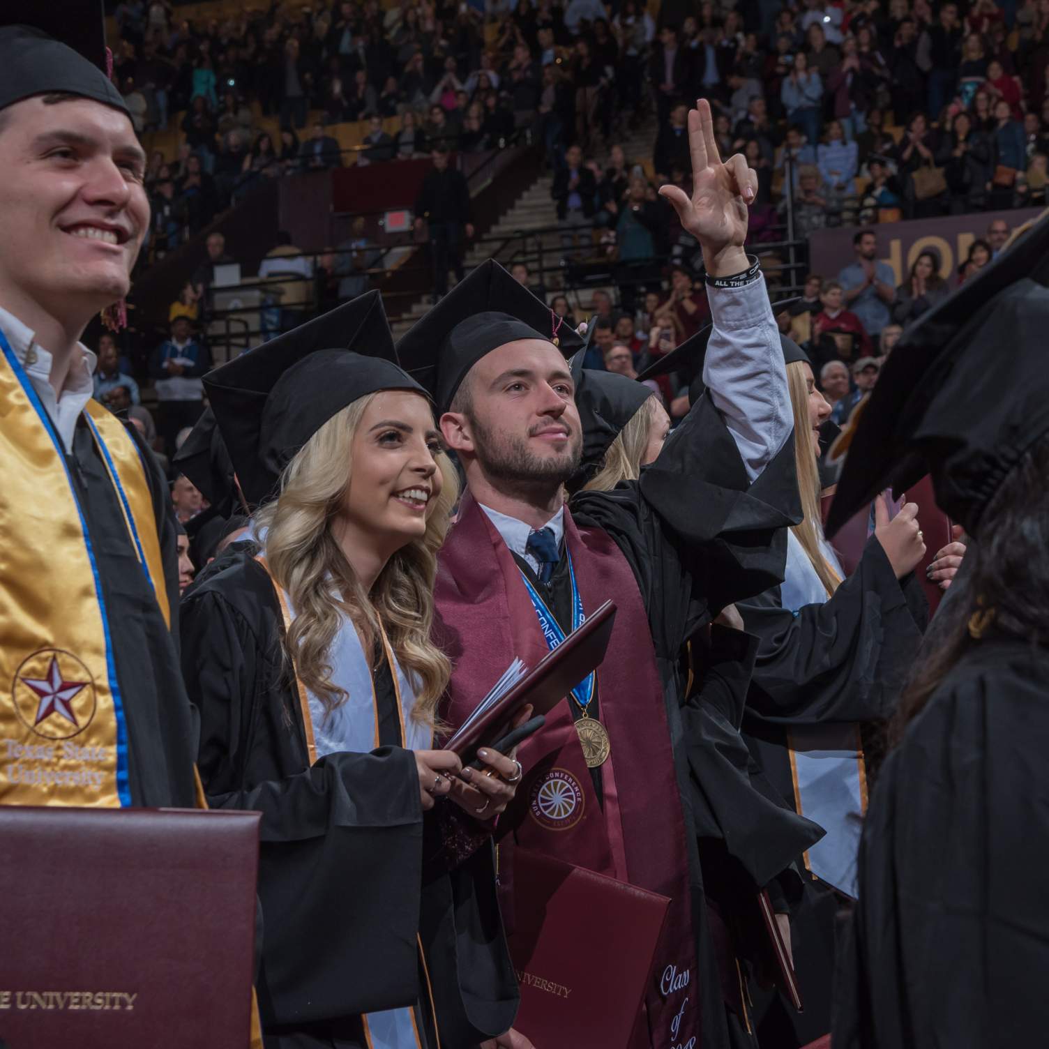 graduates holding diplomas, showing Texas State hand-sign