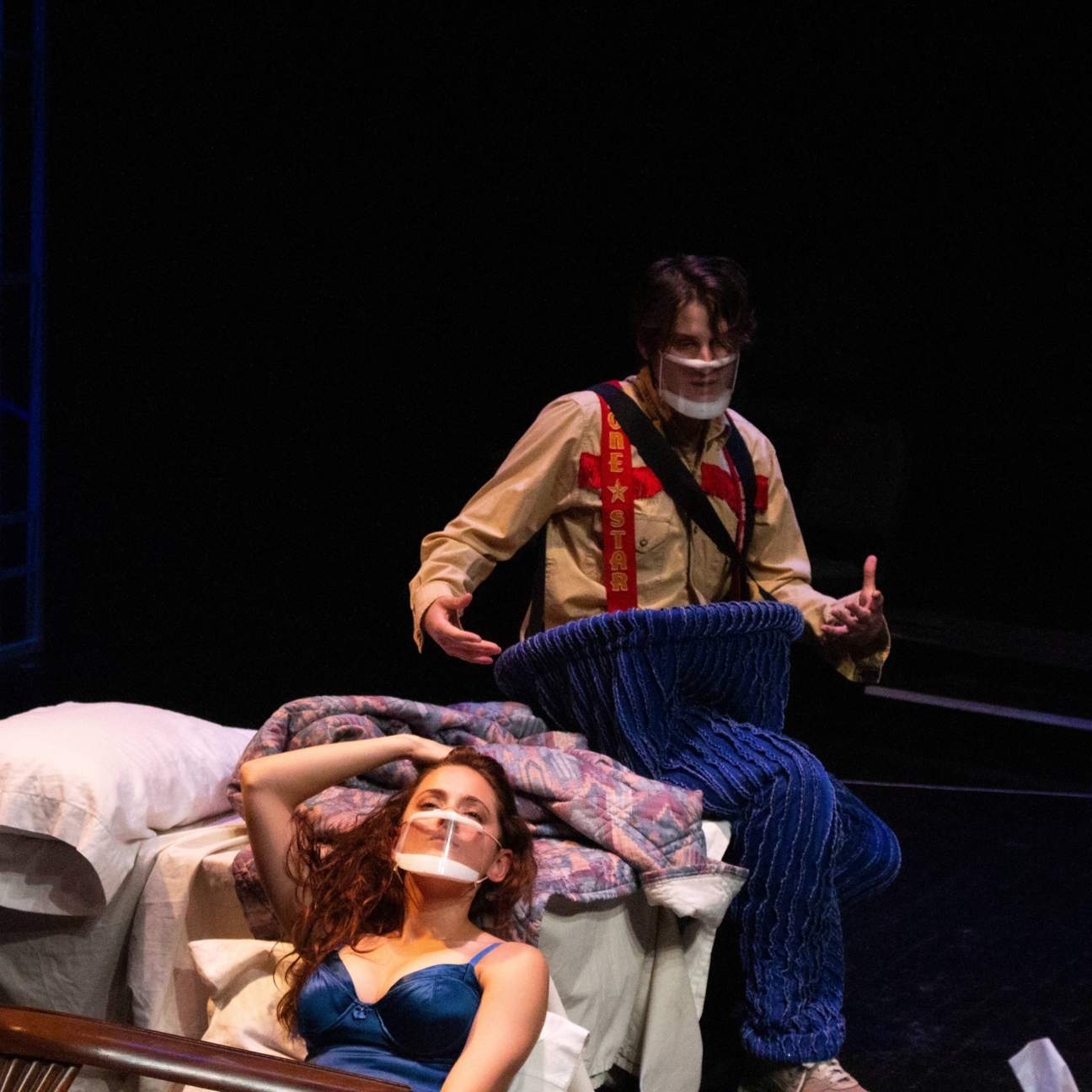 A woman lounges on a bed with one arm above her head, facing away from a man who sits on the bed behind her. The man behind her is wearing oversized pants, suspenders, and other remnants of his costume. 