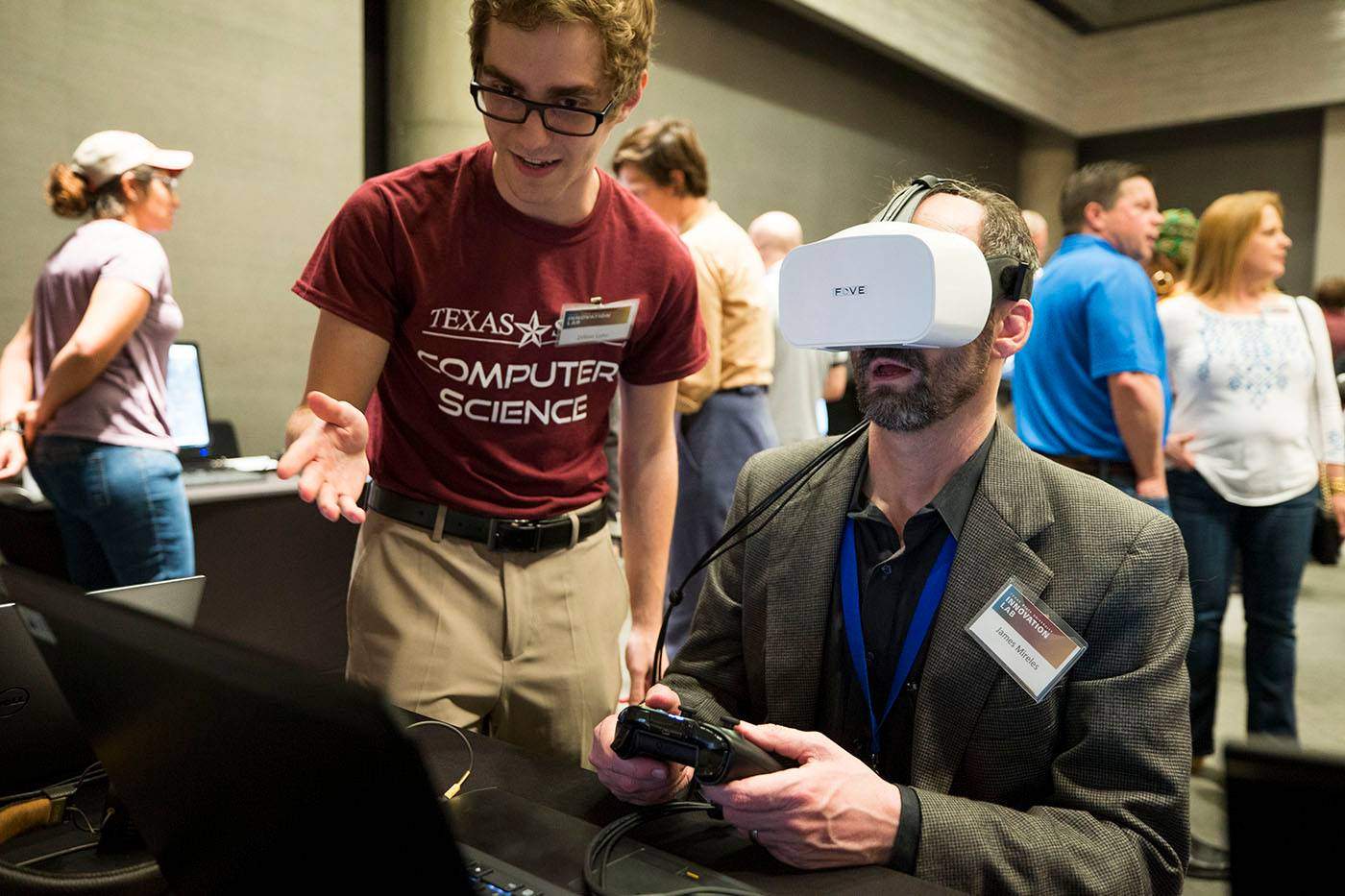 Photo of student at Innovation Lab event demonstrating virtual reality as a seated visitor wears the virtual reality goggles
