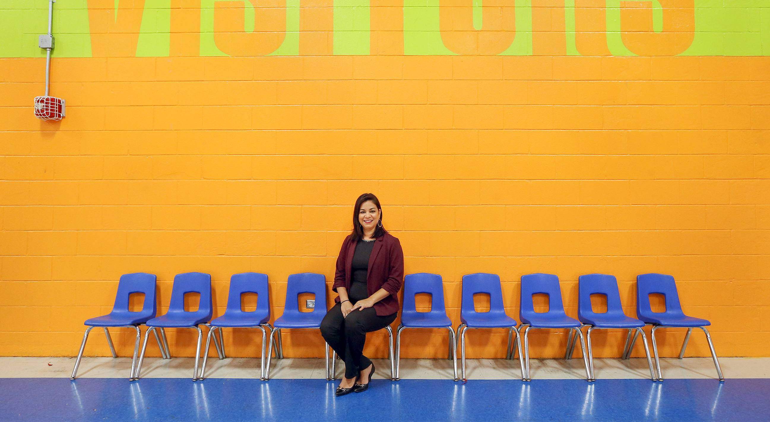 Gonzalez-Alcantar sits in the middle of a row of blue classroom chairs against a bright orange brick wall