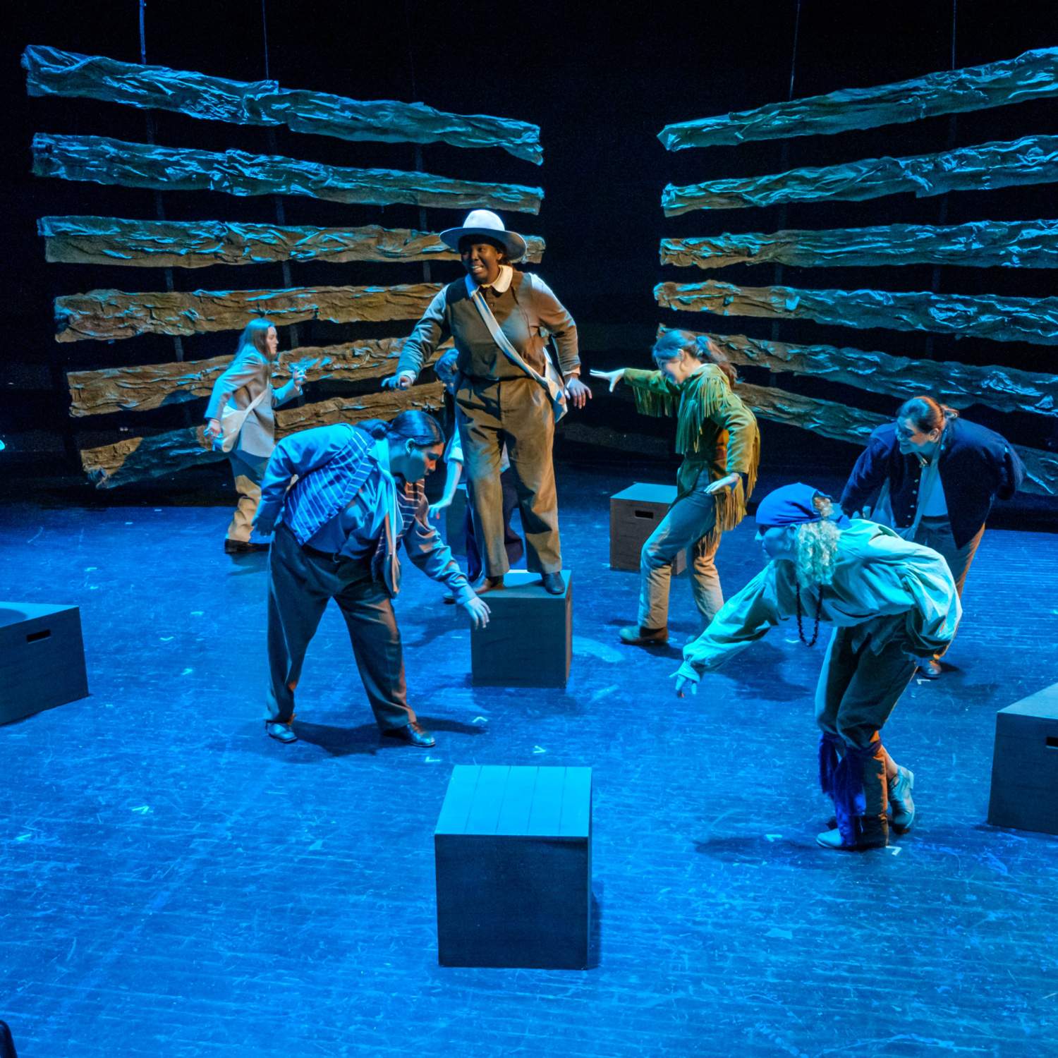 One woman in frontier clothing stands on a crate above the rest. The stage is washed in blue light as several other women crouch and crawl around her on the ground.