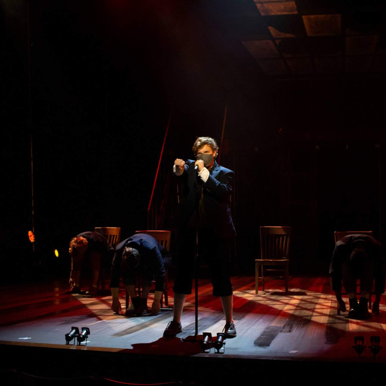A young man in a sweater stands at his microphone stand, singing aggressively while pointing out at the audience. Behind him, his classmates sit in their chairs with their heads on their knees. Red light washes the stage.