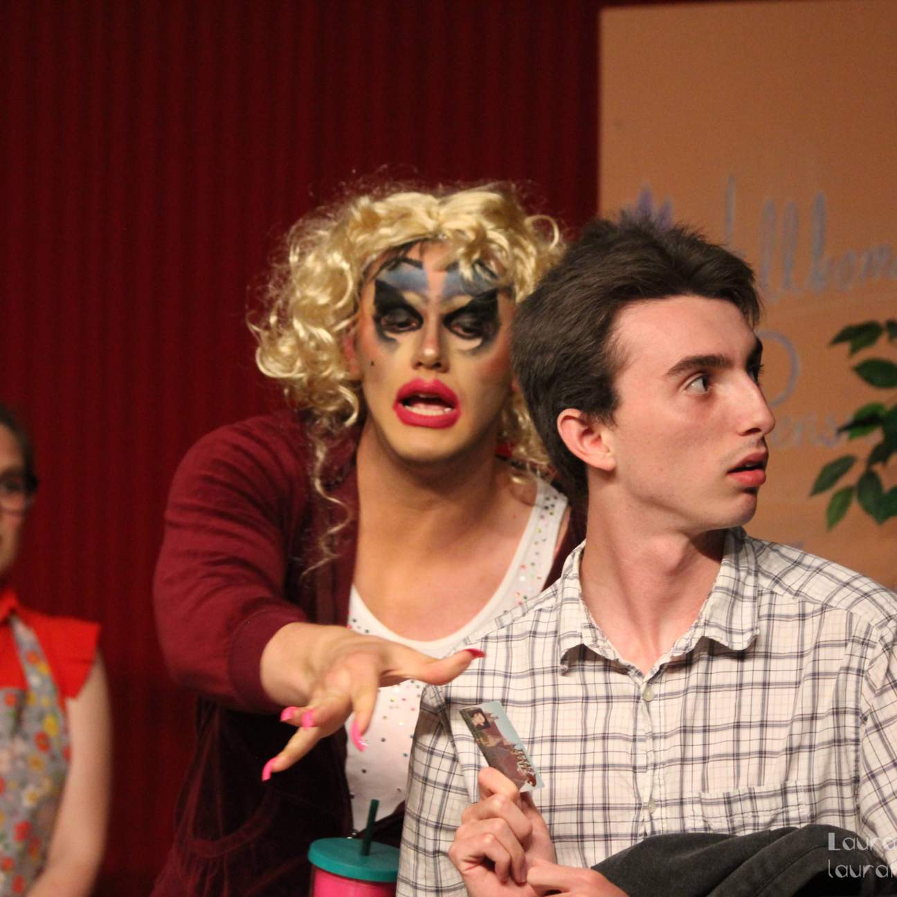 A man in drag reaches from behind another man, while a woman in an apron is in the background