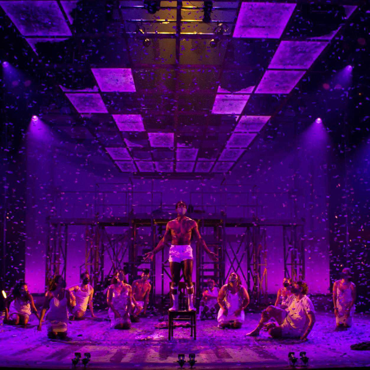 The stage is washed in purple light as purple petals rain down on the cast and fill the entire stage. The whole cast is on stage in their underclothes sitting looking up at the petals in joy and wonder, excluding a young man who is standing on a wooden chair above the rest with his arms spread wide.