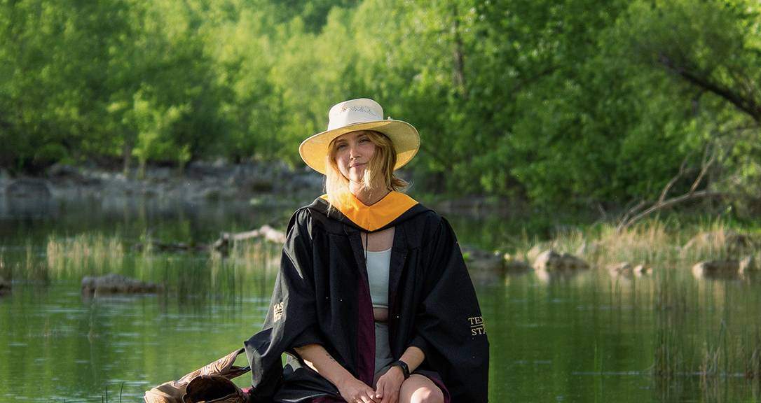 woman in graduation regalia sitting on river rock