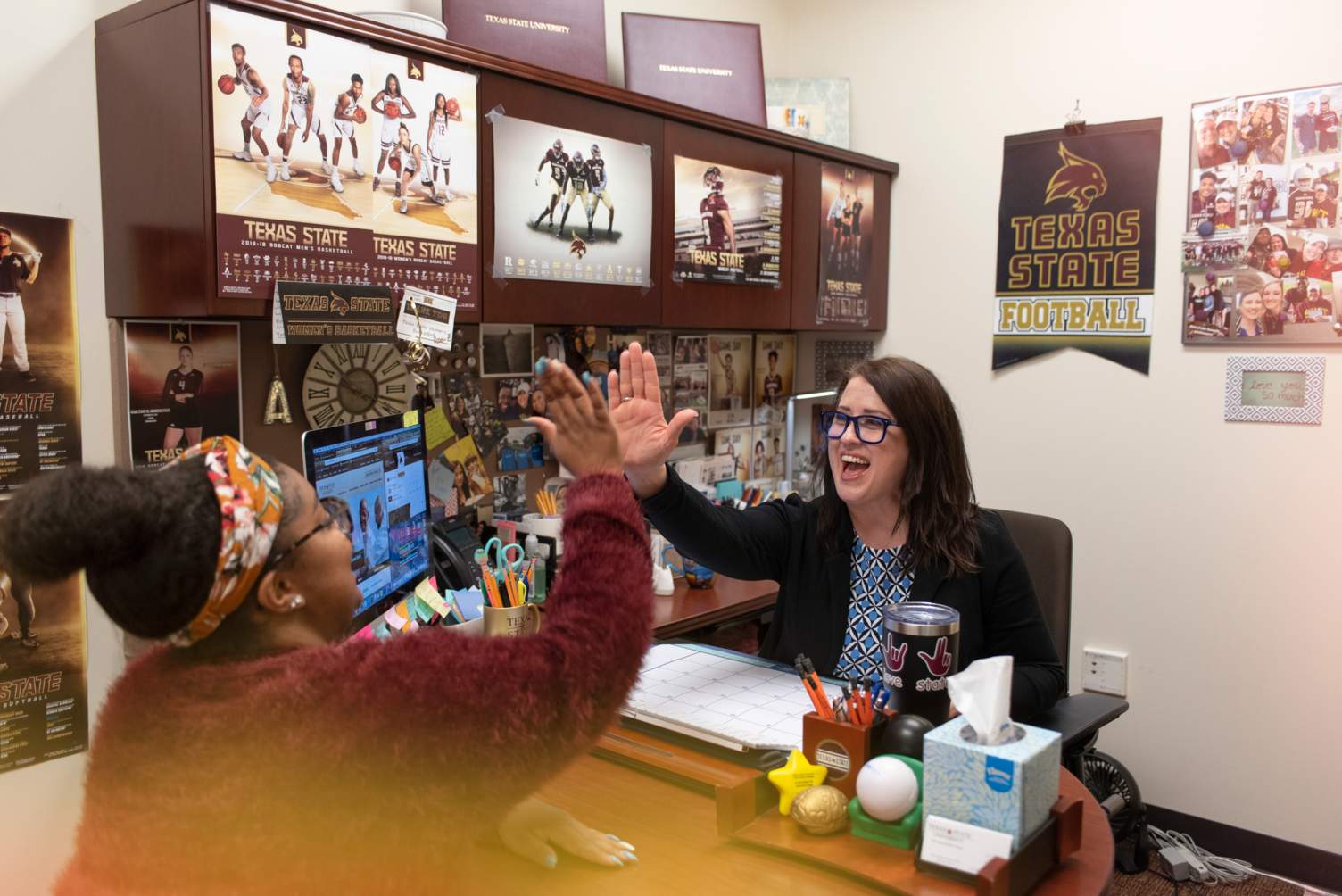 Two women in an office high-fiving. Lots of Texas State memorabilia on the walls. 