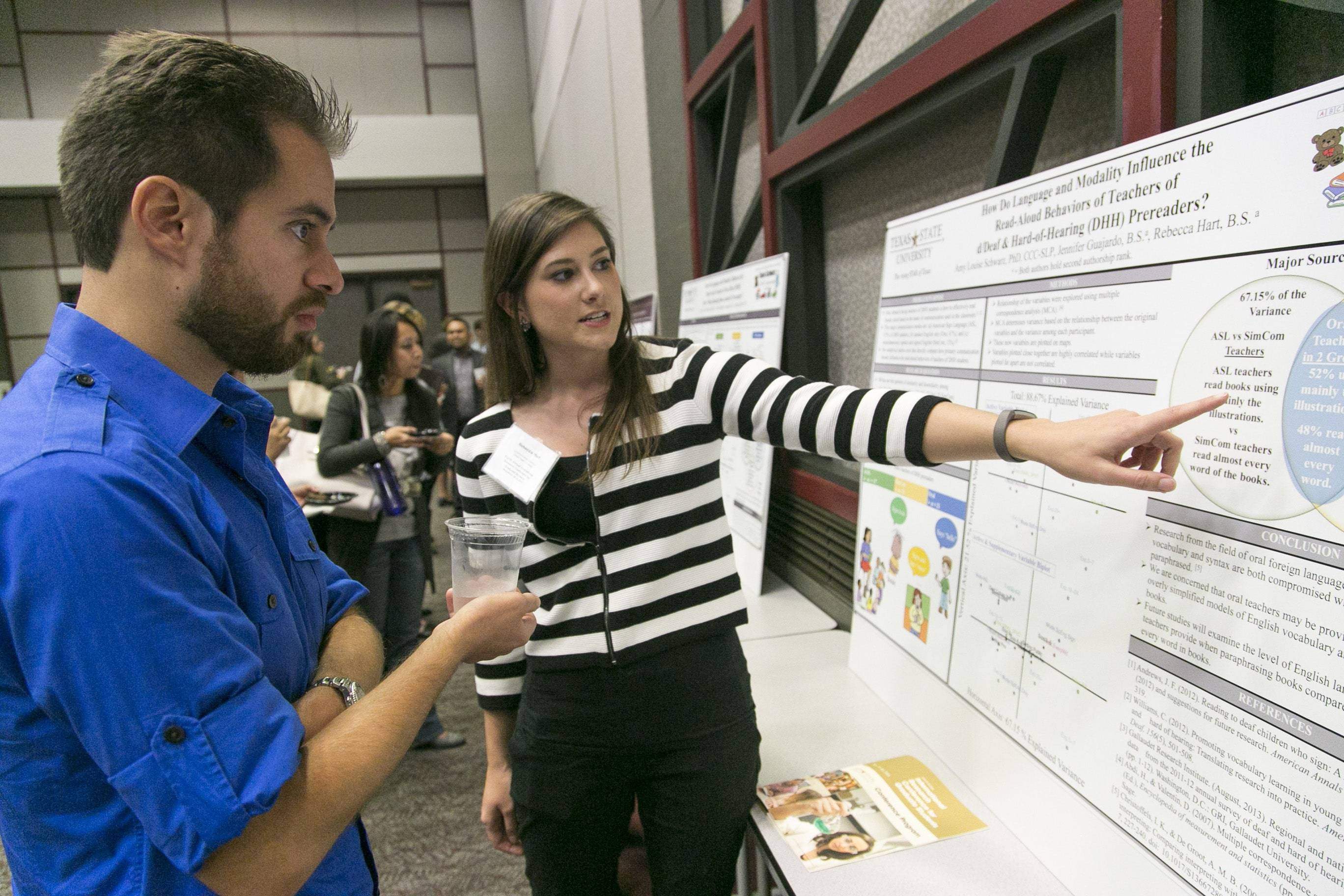 A female student points to her research poster while explaining her work to a male student.