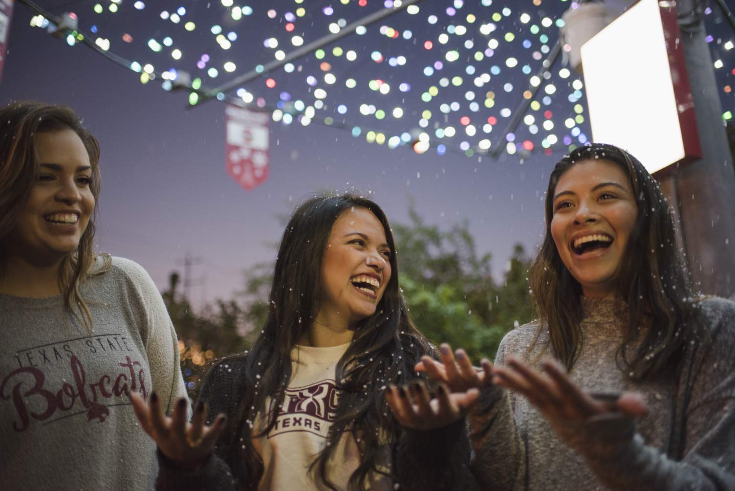 Three students laughing while fake snow falls on them. 