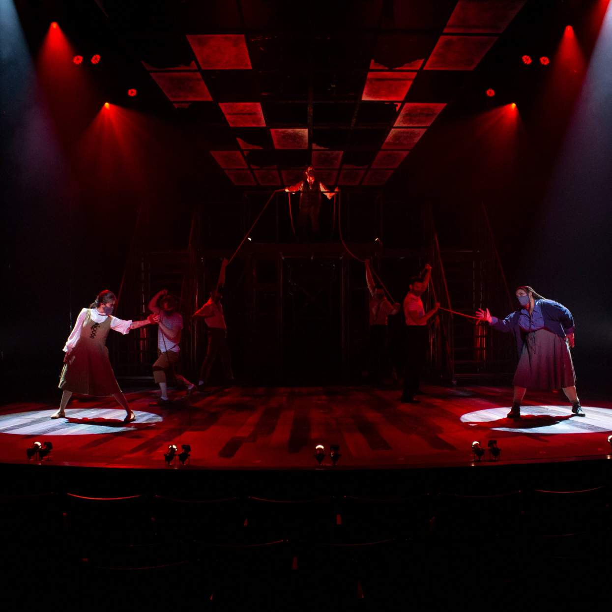 A menacing man stands on a platform above two young girls. He holds red ropes which are tied to each of the girls hands, binding them to him. Male figures, hold on to the ropes and pull on them, jerking the girls and keeping them apart from each other. The stage is washing in red light, and their are spotlights on each of the girls.