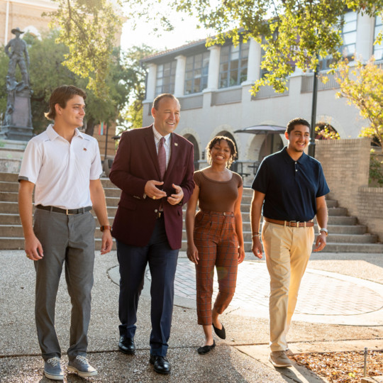 President Damphousse smiling with three student in front of Old Main and Lampasses
