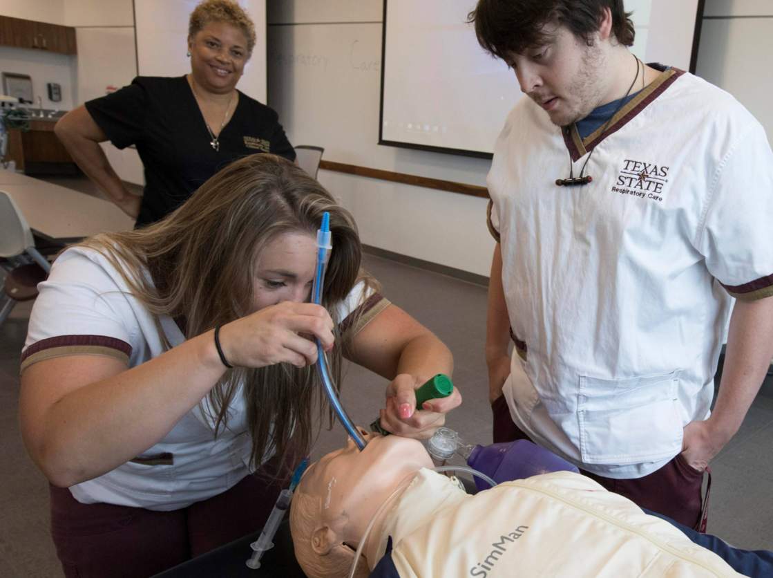 Student practicing intubation on a manikin in a classroom while two others observe
