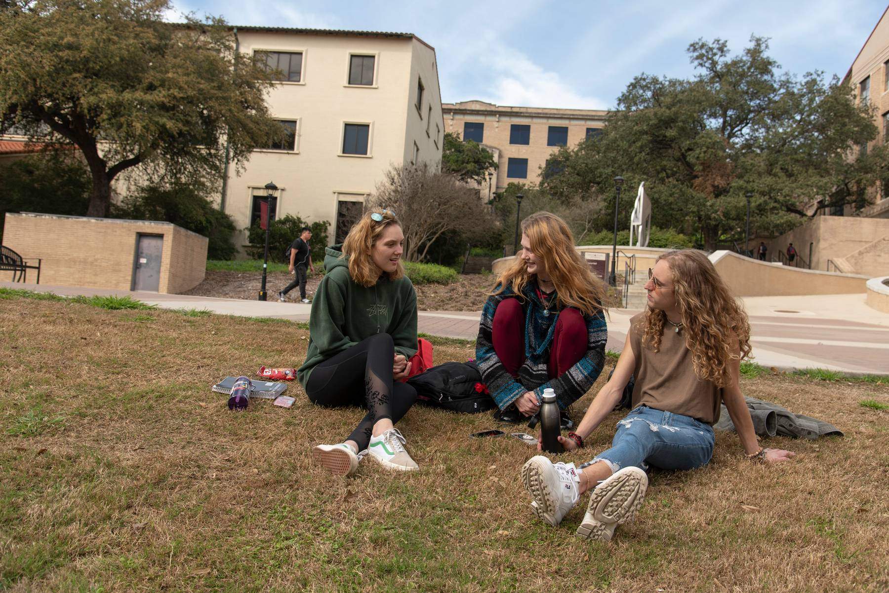 Three students sit on a grassy hill talking on campus.