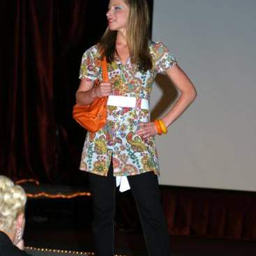 Student wearing bright colored paisley print long shirt, white belt, jeans, a chunky orange & a chunky yellow bracelets, jeans, orange purse and high-heels.