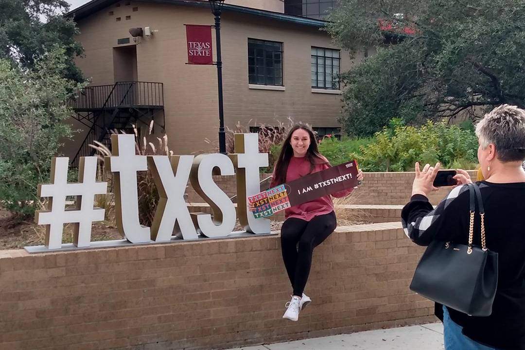 Parent taking photo of student posing with the #txst sign and holding an admitted student banner.
