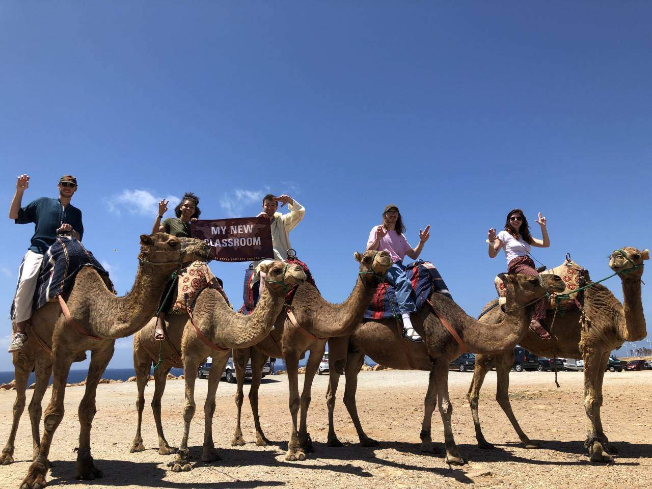 Photo of students riding camels and holding a banner reading "My new classroom"