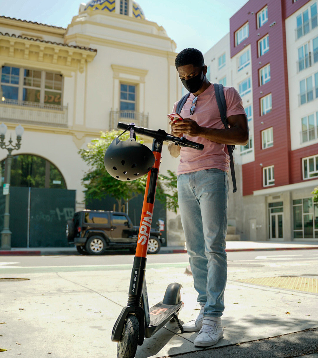 Student wearing a mask, using a smart phone while standing next to a Spin scooter