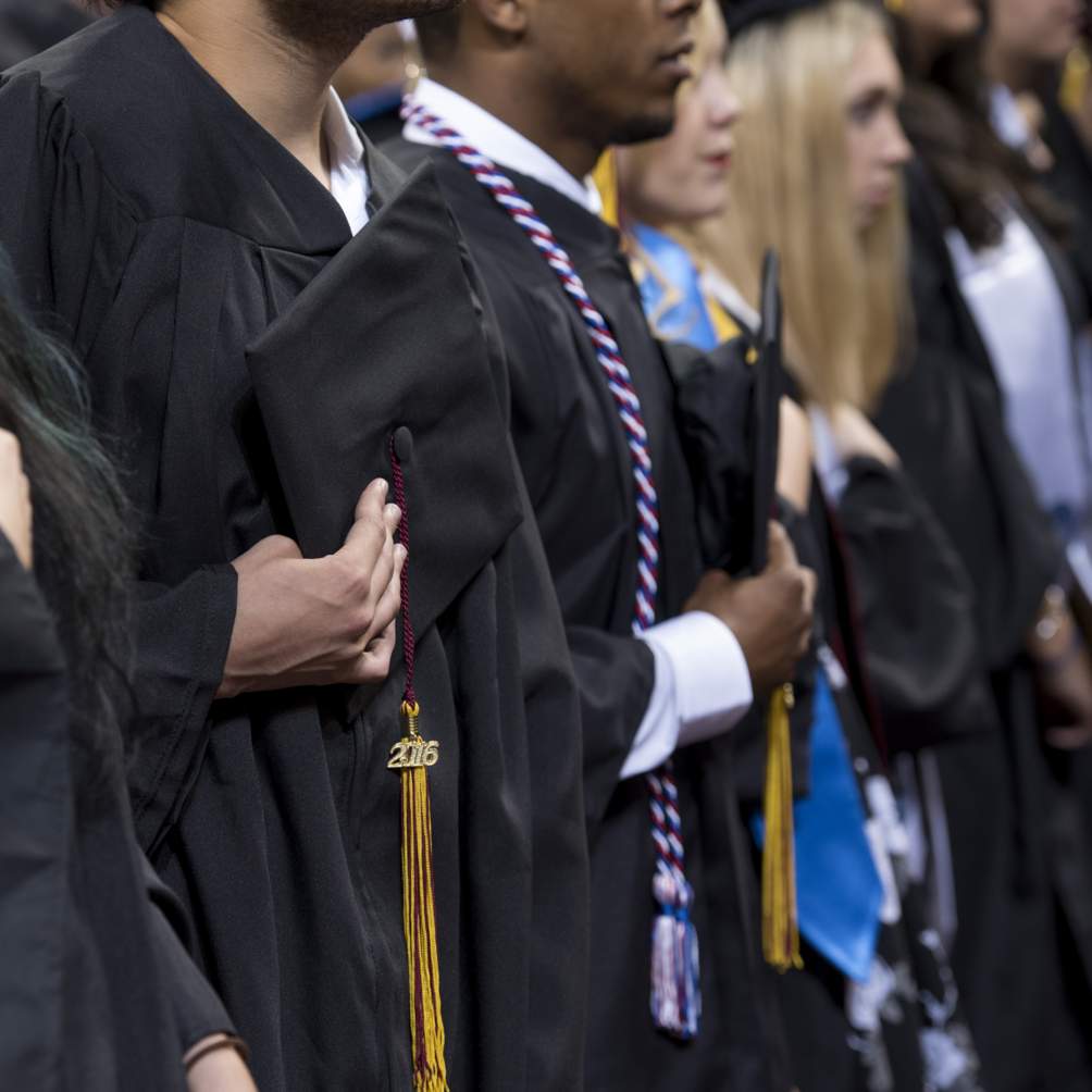 A veteran, indicated by the red, white and blue cords, removes his graduation cap as he stands with his fellow graduates for the national anthem at a Summer 2016 commencement ceremony.