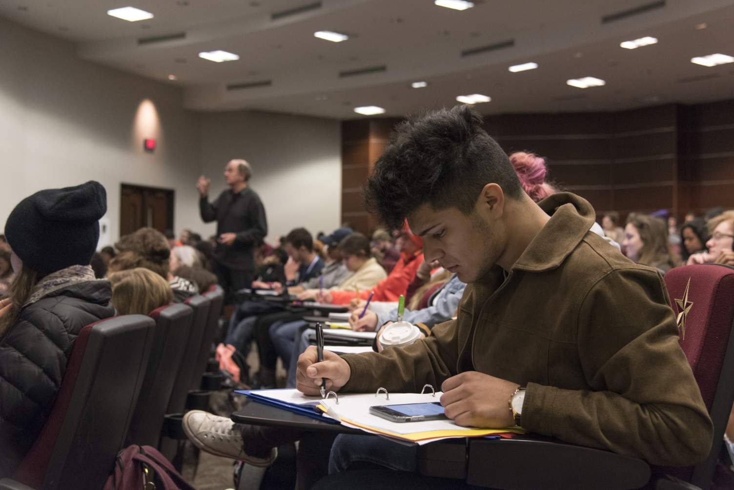 A hispanic student takes notes at a lecture in the Alkek Teaching Theatre.