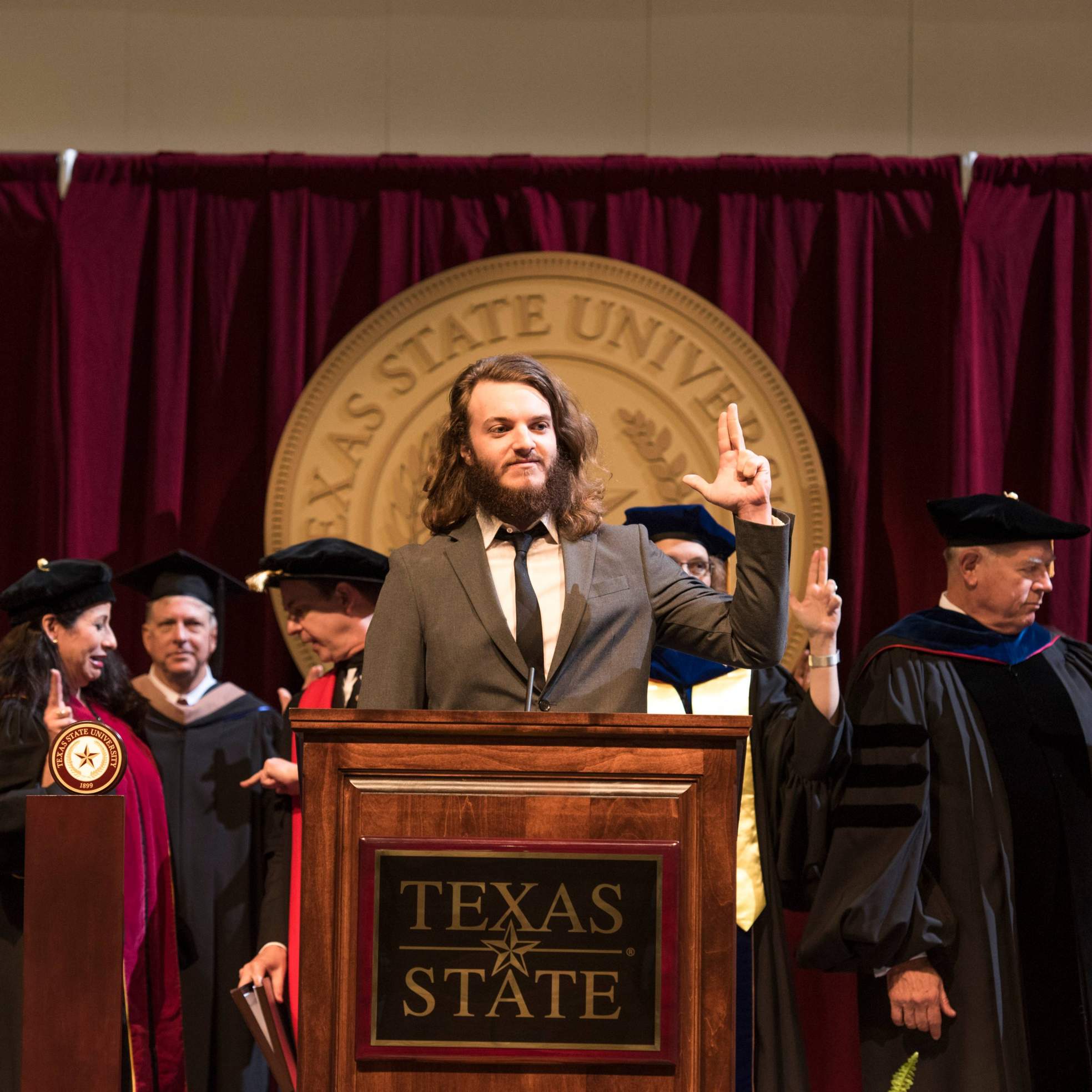 Mr. Zackerry L. Wiggs leads the singing of the Texas State Alma Mater with the state of Texas hand symbol at the closing of a Summer 2016 commencement ceremony.