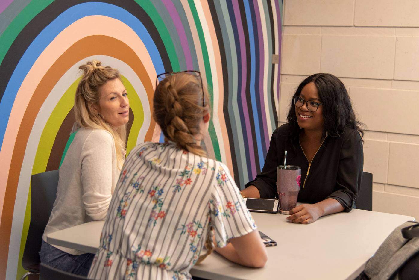 three women students chat at a small table next to a colorfully painted wall