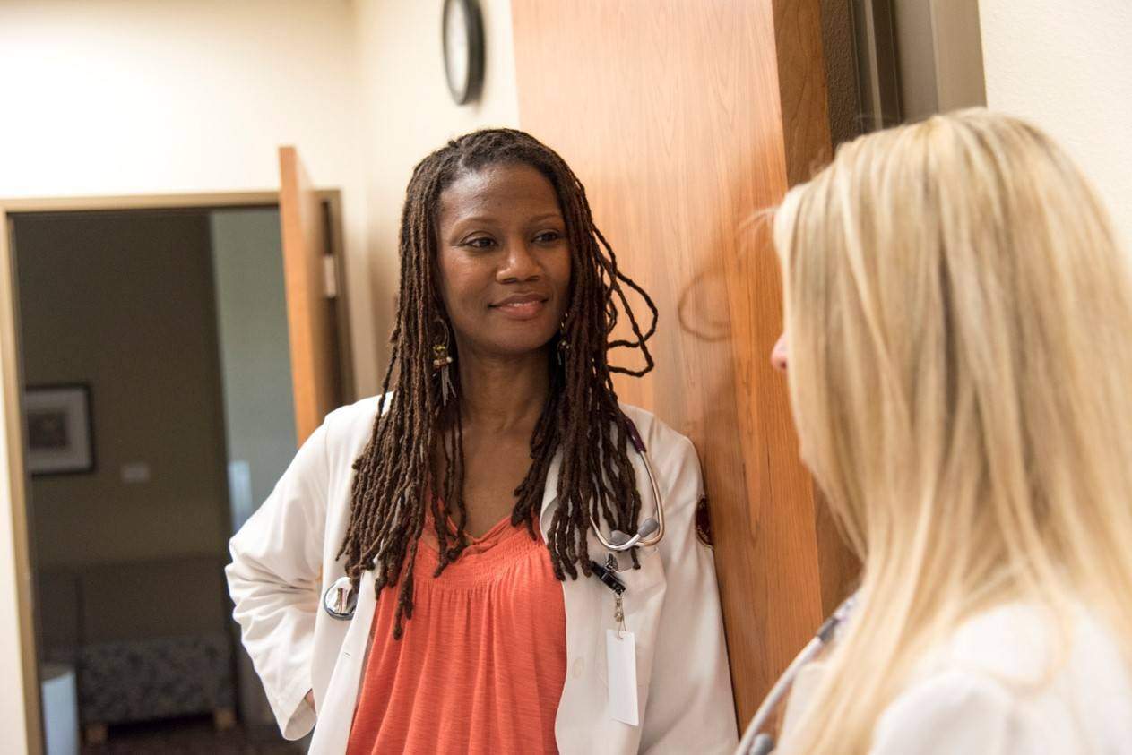 Black woman with braids in a white lab coat spaks to a person with long blonde hair.