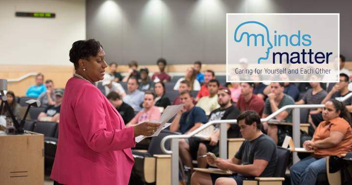 Professor lecturing to her class with Minds Matter logo. “minds matter" text is in two different shades of blue. The initial “m” spreads into an outline of a human head. The “m” in the head depicts a brain.