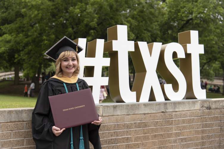 graduate next to #TXST sign