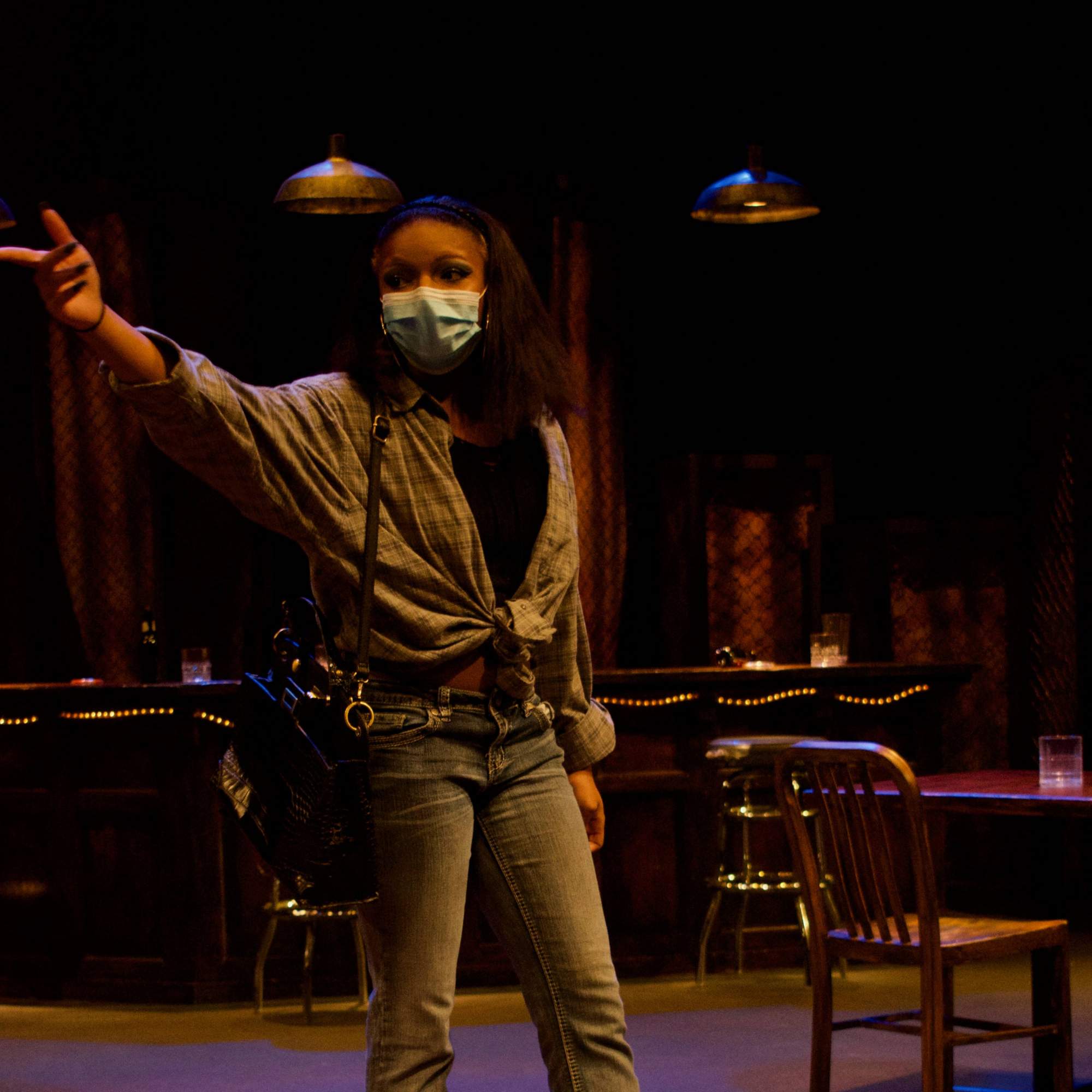 A woman in a tied up plaid shirt, jeans, and boots stands in the bar while pointing to something out of frame. 