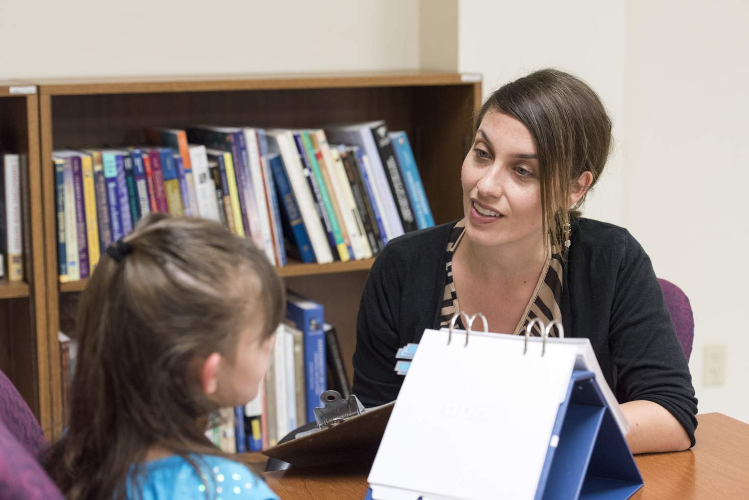 a school psychologists conducts an assessment with a young student, the school psychologist is writing on a clipboard and a small flip chart is situated on the table between the pair