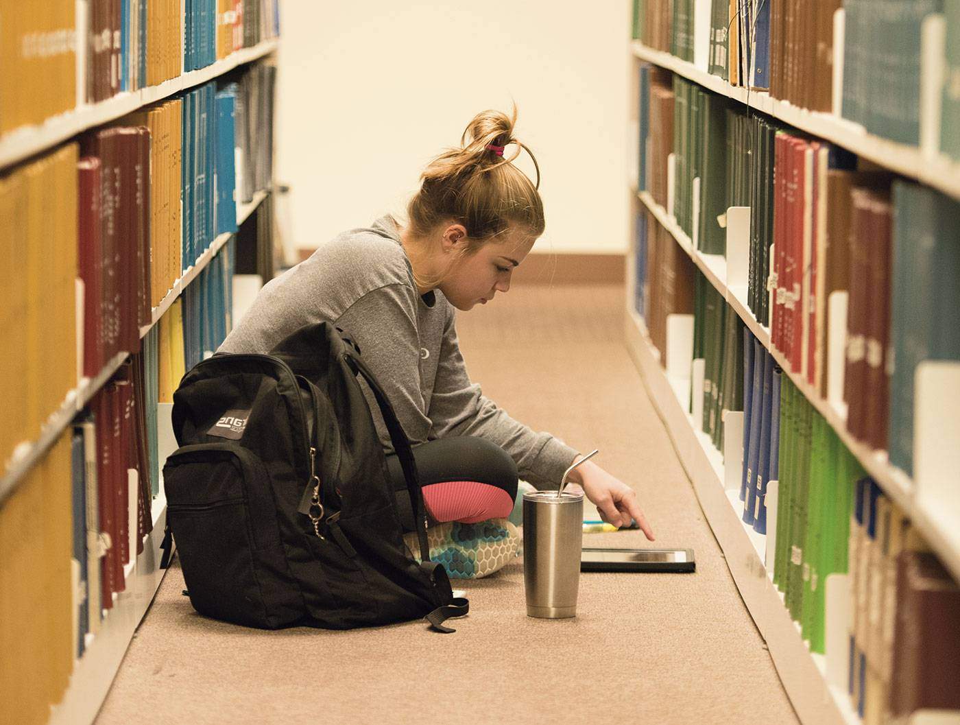 a female student sits on the floor of the library in between two bookshelves to study
