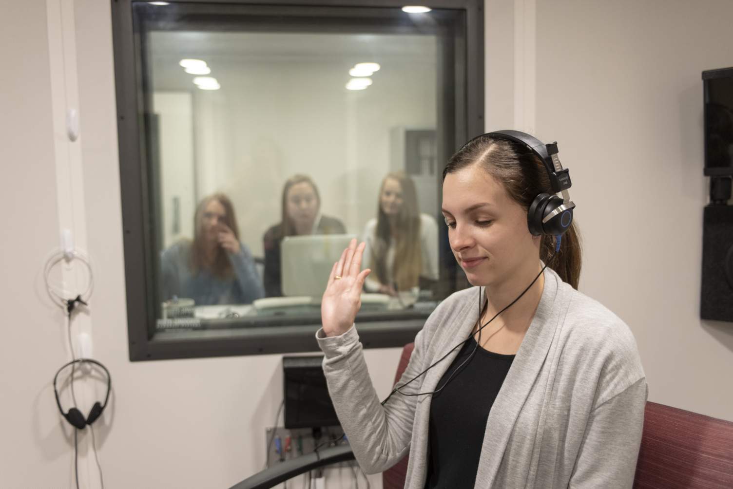 Subject in hearing clinic wearing headphones with their eyes closed and a hand raised, and three students observing through a window from another room