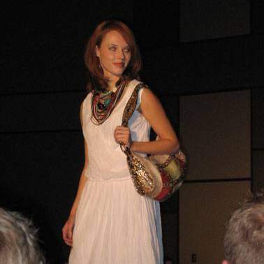 Student/model in white dress, wearing necklaces, carrying colorful purse walking down the runway. 