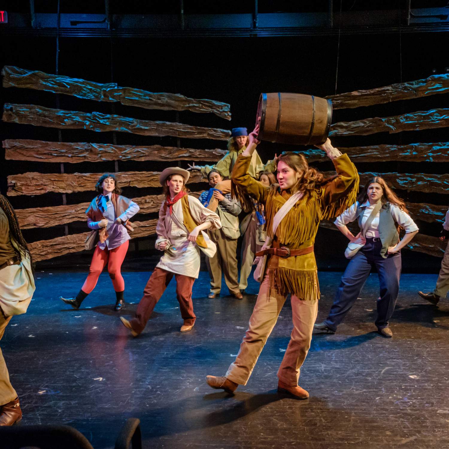 One woman in frontier clothing holds a barrel over her head while several other woman dance behind her, lifting one woman into the air. 
