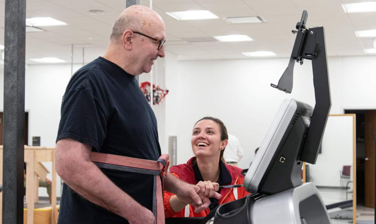 A young physical therapist helping an older man walk on a treadmill with support