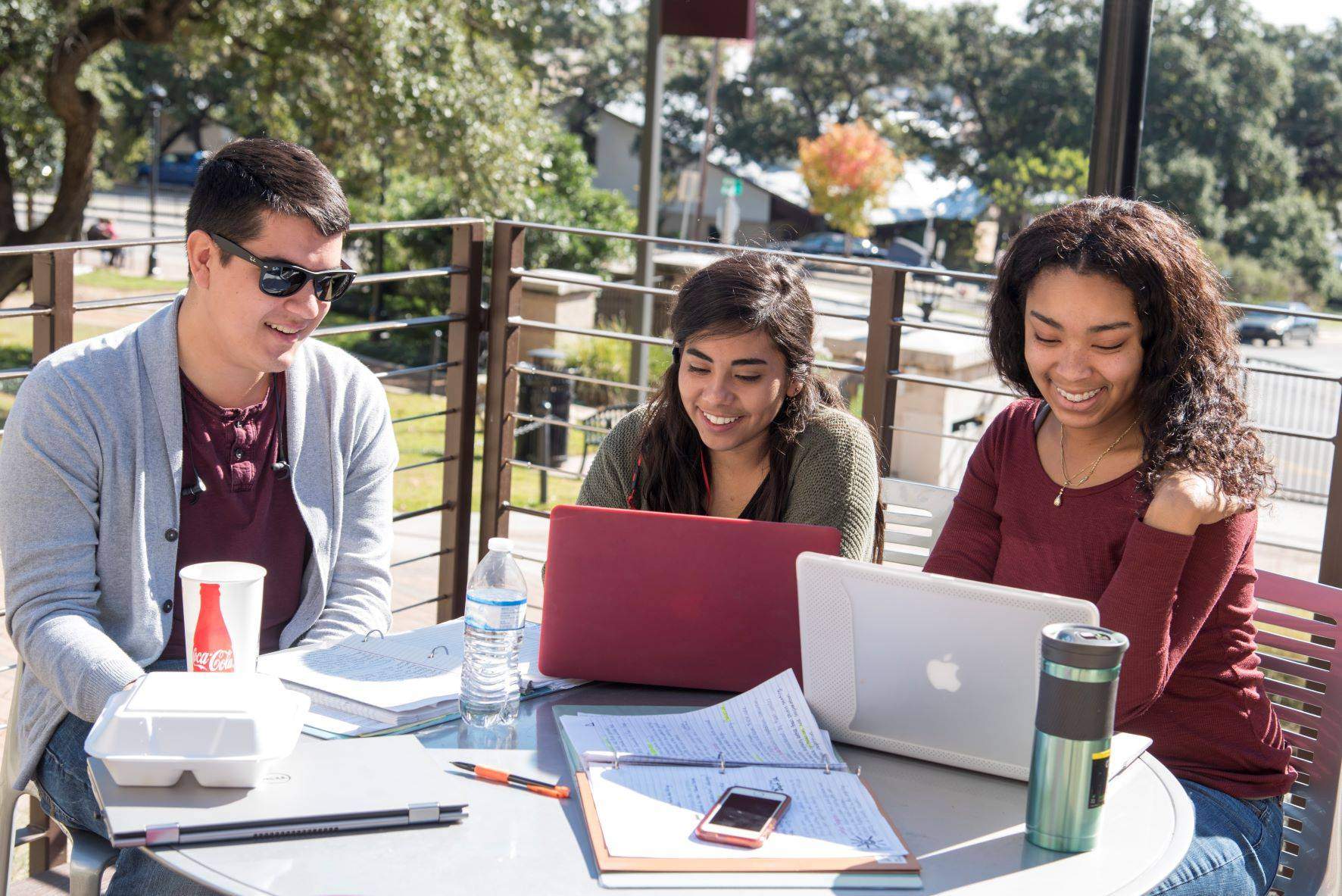 three students sitting at a table outside with laptops
