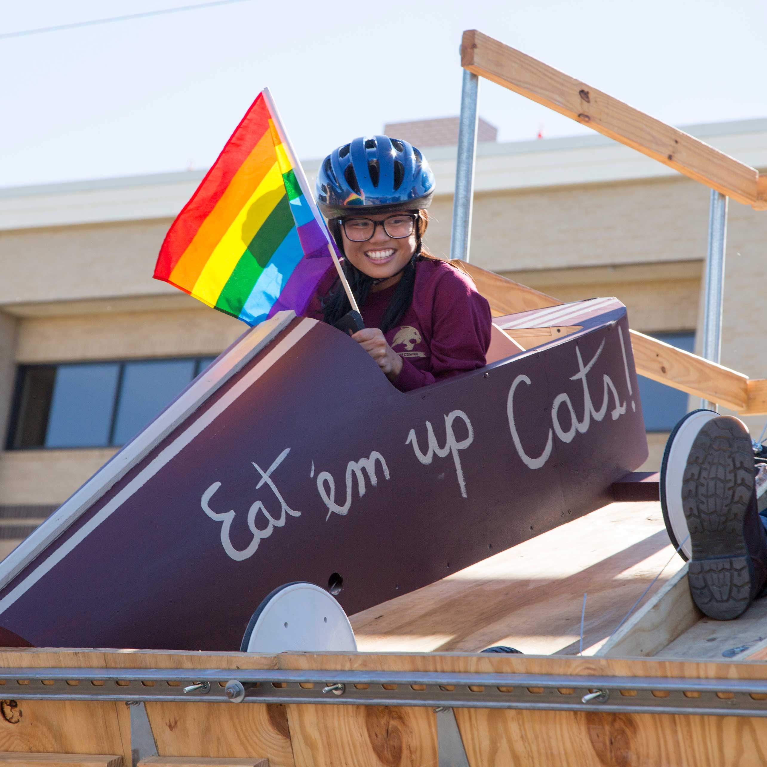 a young female student hold a rainbow flag as she prepares to take off in the homecoming derby race