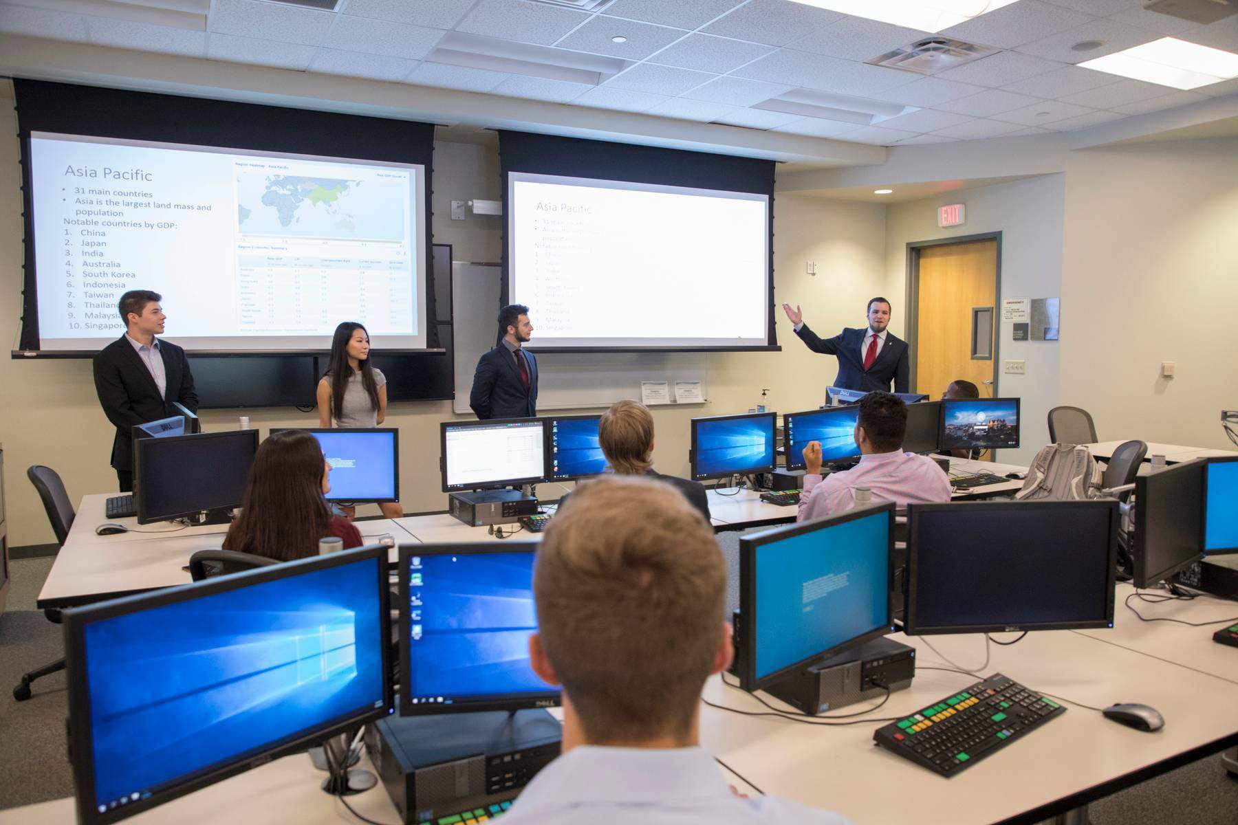 photo of students in a computer lab, multiple students giving presentation at front of room
