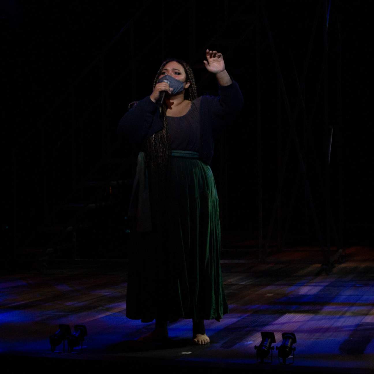 A young girl in a long loose skirt, peasant shirt and knit jacket stands barefoot singing into her microphone, her other arm is raised above her head and she looking longingly into the audience
