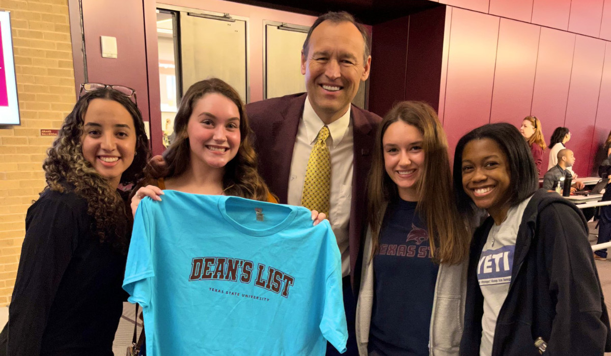 President Damphousse smiling with students while one holds up a t-shirt that says: Deans List