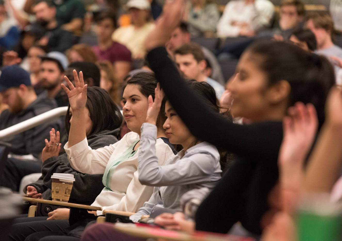 Students with raised hands sitting in a large lecture hall